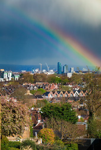 Panoramic view across the city of London, taking in huge residential districts, with the ultra modern architecture of the skyscrapers of downtown visible on the horizon. There has been a rain shower and a rainbow is vivid in the sky.