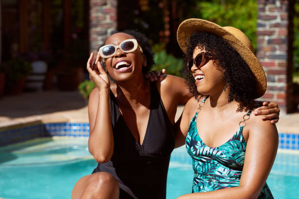 Two Black friends laugh and joke sitting by swimming pool at home, summer fun