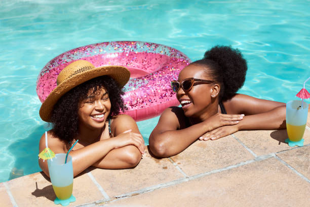 Two Black friends lean on the side of the pool, relaxing in water with mocktails