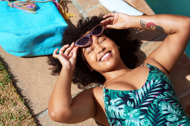 Overhead shot of young Black woman lying next to pool with purse, sunscreen