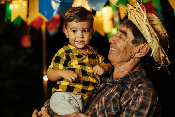 Photo of Senior man and his baby grandson celebrating the Brazilian Festa Junina. Portrait of grandfather and his grandson wearing typical clothes and a straw hat during the traditional June festival in Brazil.