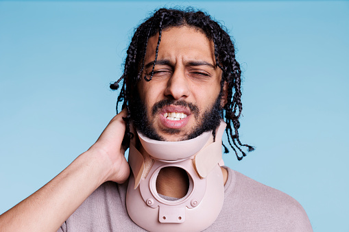 Arab man wearing cervical collar and grimacing in pain while touching neck. Injured young person with painful facial expression wearing brace, recovering from spinal trauma