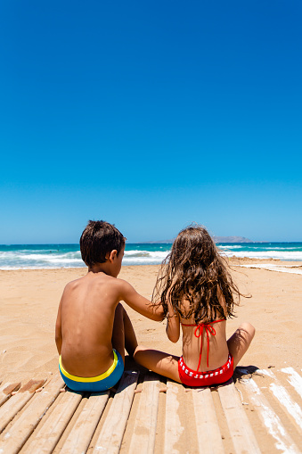 Girl and boy sitting on the sand at the beach on their summer vacation