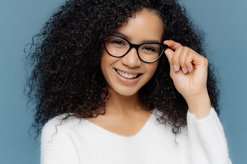 Headshot of optimisitc young African American woman with crisp hair, holds hand on frame of glasses, wears white sweater, being in good mood after successful day, isolated over blue background.