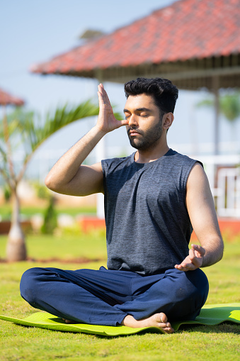 full shot of Young man doing nostril breathing exercise or pranayama yoga by eyes closed at park - concept of Zen, peaceful and self care