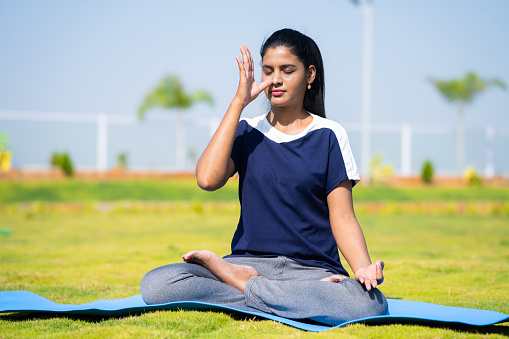 Young girl doing nostril breathing exercise or pranayama yoga with closed eyes at park - concept of self care, mindfulness and active morning routine