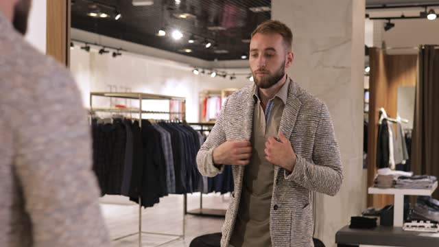 Shopping and fashion concept - Young bearded man choosing and trying jacket on in mall or clothing store.