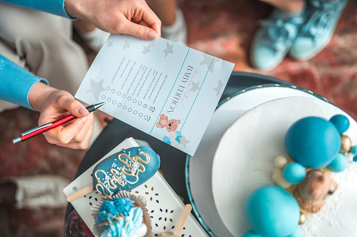 A hand is holding a paper with an illustration and space for guests to participate in a baby shower activity. The photo features a colorful and festive backdrop of decorations, as well as beautiful desserts.