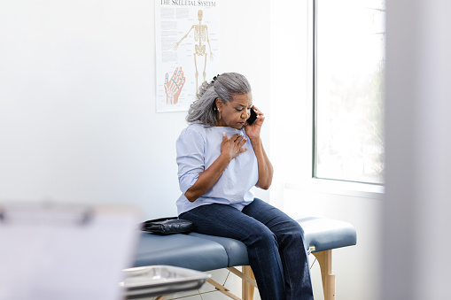 The mature woman sits in the doctor's office and holds her chest while talking to her friend who is telling her some difficult news.