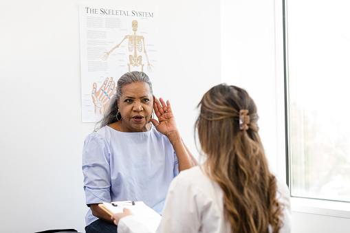 While the doctor is talking, the mature woman places her hand to her ear to let her know that she can't hear very well.