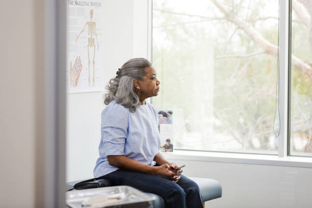 Mature woman holds her mobile phone and waits for the doctor The mature woman sits on the examination table and holds her mobile phone as she waits for her doctor to enter the room. waiting stock pictures, royalty-free photos & images