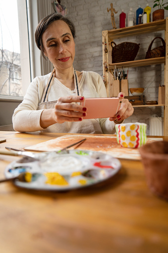 mature woman photographing her pottery bowl in workshop. Craftsperson painting a bowl made of clay in art studio