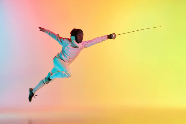 istock Long attack. Young man, male fencer with sword practicing in fencing over gradient pink-yellow background in neon light. Sportsman shows fencing technique 1483065100