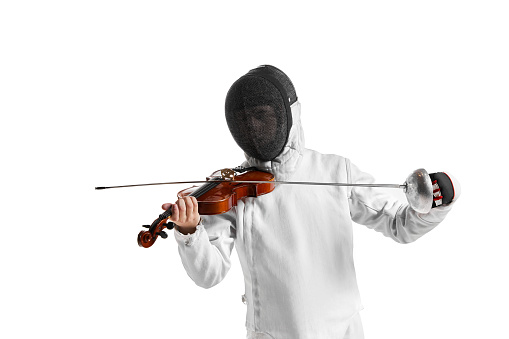 Young man, male fencer in fencing costume and protective mask helmet playing violin over white background. Concept of sport, music, hobbies. Copyspace for ad.