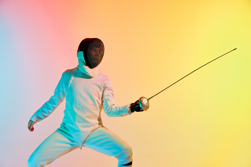 Young man, male fencer with sword practicing in fencing over gradient pink-yellow background in neon light. Sportsman shows fencing technique. Copyspace for ad.