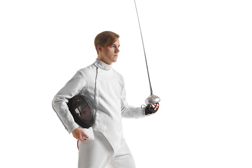 Young man, male fencer in fencing costume mask standing with sword over white studio background. Sport, motivation, professional skills. Copyspace for ad, text.