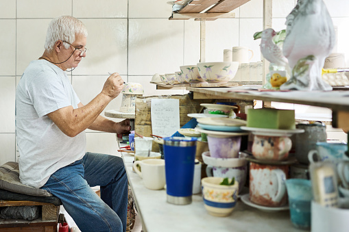 Senior male worker painting floral design on a bowl at a table in a ceramics manufacturing workshop