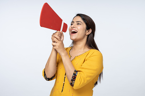 Portrait of young woman screaming in megaphone, announcing important information on white background