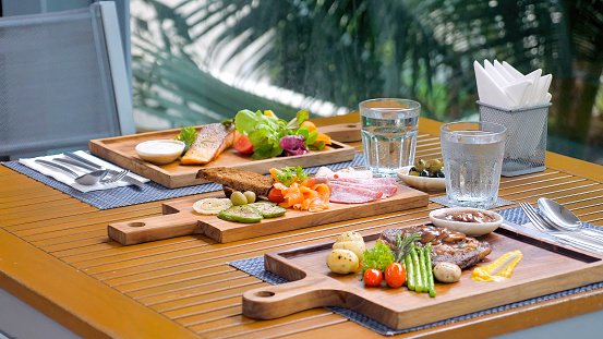 Outdoor restaurant table displays variety of meat cuisine, including grilled beefsteak, smoked salmon, and vegetable omelets. A gourmet meal for any summer holiday.