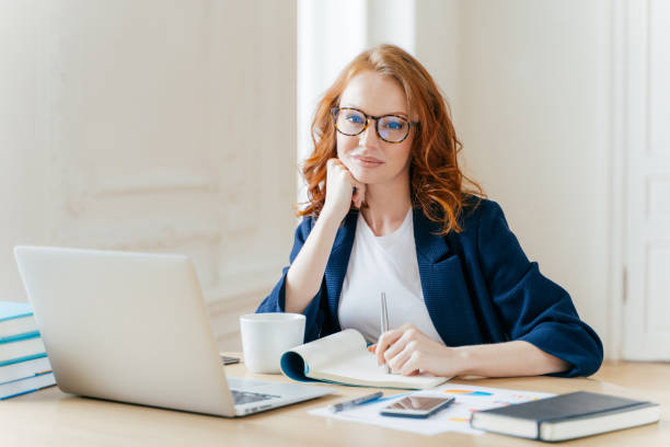 istock Redhead curly woman office worker analyzes data, makes accounting report, poses in coworking office, improves online sales, drinks coffee, wears spectacles and formal wear. Female employee indoor 1483074464