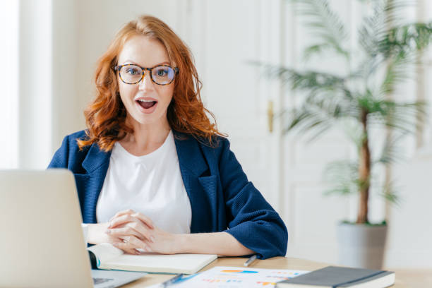 istock Surprised ginger female employee has job, develops new business srategy, poses in front of opened laptop computer, poses in workspace against office interior. Woman software developer at work. 1483074471