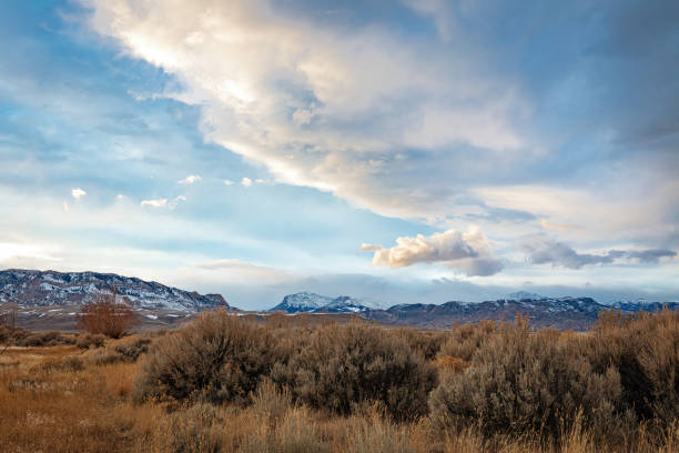 Panoramic landscape at sunset of cumulus clouds over the high desert wilderness of northwest Wyoming in winter with sagebrush in the foreground and snow capped mountains in the midddle ground just below the sky line.