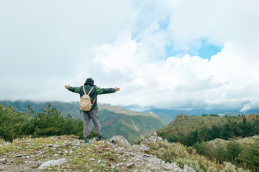 Traveler man hiking in mountain forest, standing on the stone with raised up arms against panoramic mountain valley.
