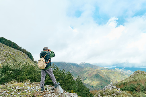 Traveler man hiking in mountain forest, standing on the stone looking with binoculars panoramic mountain valley.