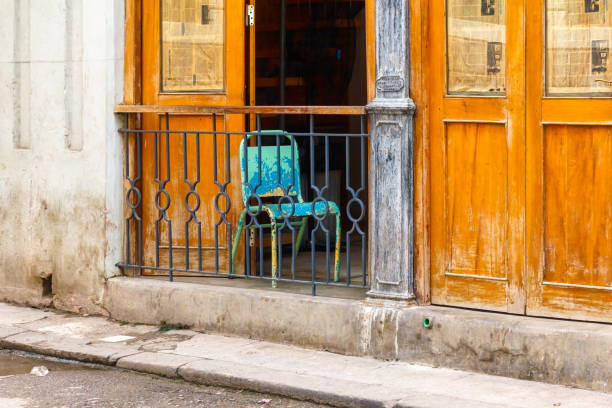 Havana, Cuba - April 10, 2023: Close-up of two wooden doors on a building. One of the doors is open, with a metal railing at the front and a blue chair behind the railing.