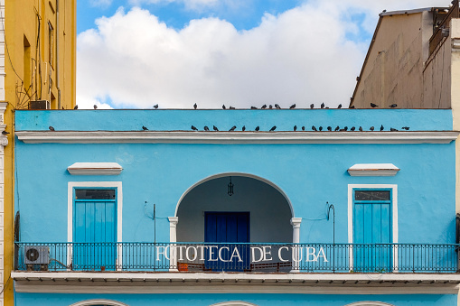 Havana, Cuba - April 11, 2023: Closeup facade of a blue-story building with a balcony and an archway leading to an entrance. There is a metal railing on the balcony with the words 