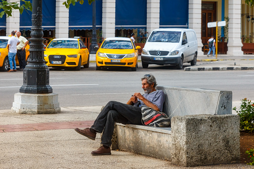 Havana, Cuba - April 11, 2023: A man is seated on a concrete chair in a public park adjacent to a street. A building with columns is visible in the background, and other incidental people and parked vehicles can be seen in front of the building.