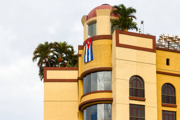 Havana, Cuba - April 12, 2023: Low-angle facade of a building with glass windows, a dome, and palm trees on the roof. A Cuban flag hangs from the top, and no people are on the scene.