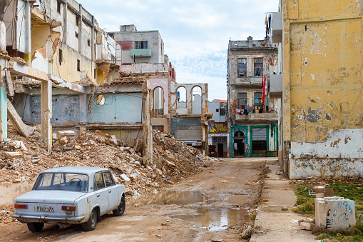 Havana, Cuba - April 12, 2023: A street with a parked car beside a damaged-story building, surrounded by rubble. On the left side of the road are other story buildings with faded and worn paint on their walls.