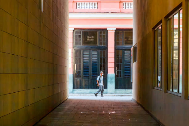 Havana, Cuba - April 12, 2023: A man wearing a facemask and carrying a knapsack walking in front of a building with a glass front in the background. In the foreground is a passage with high walls on opposite sides, with one side having glass windows.