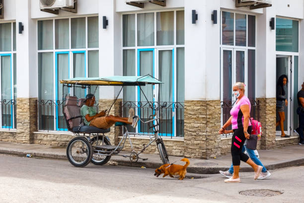 Havana, Cuba - April 14, 2023: Man sitting in a trike on a street side with his legs on the right handle grip and using a mobile phone.  A woman and a child walking a dog are in the area with a building in the background.