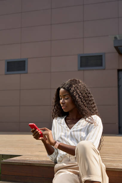 pretty young african woman model using smartphone sitting outdoors. - surfing wireless vertical outdoors lifestyles imagens e fotografias de stock