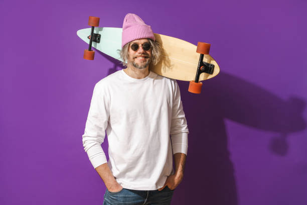 Portrait of cheerful middle aged man with longboard wearing white sweatshirt against purple background