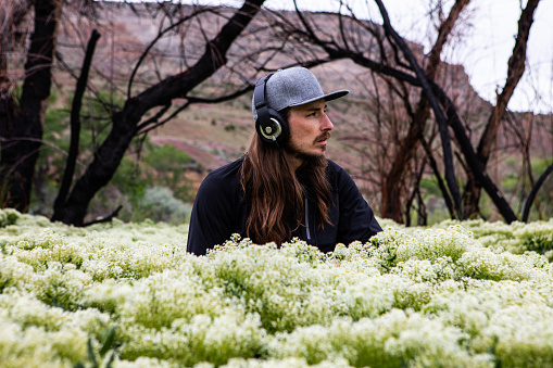 Handsome Caucasian Man with Long Hair Sitting in Flower Field Listening to Music in Headphones