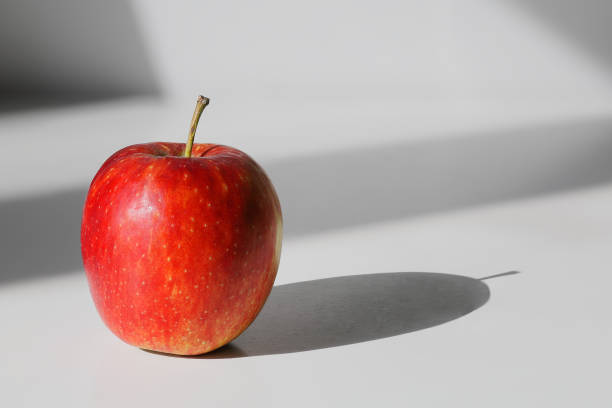 An apple on a white desk. Light from the window on a sunny day floods the table and objects leave long shadows. Selective focus.