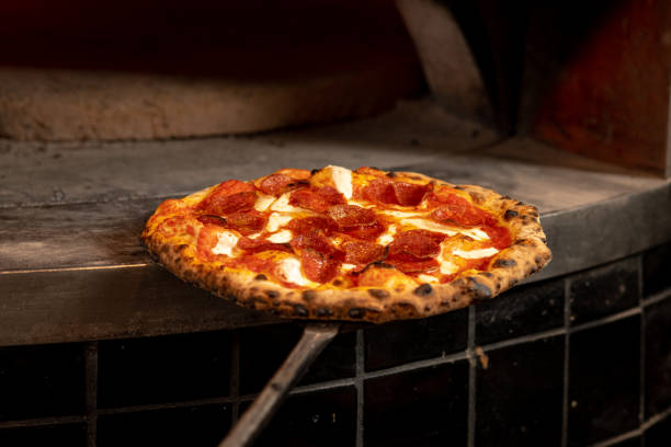 Pizzaiolo pulling a fresh pepperoni pizza from a hot pizza furnace.