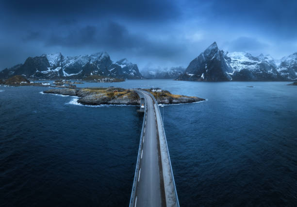 Aerial view of bridge, island with rorbu, sea, snowy mountains in fog, blue cloudy sky at twilight in spring. Dramatic landscape. Top drone view of road. Hamnoy village, Lofoten islands, Norway stock photo