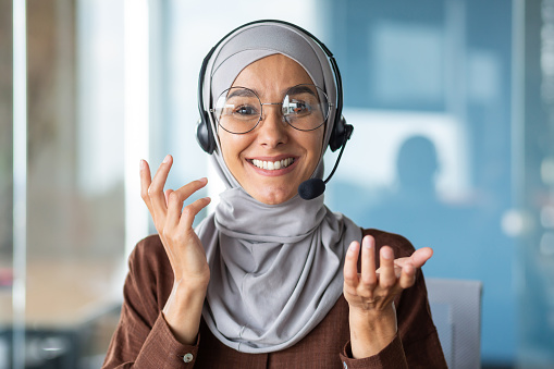 Online customer support worker consults a client remotely, a woman in a hijab with a headset for a video call talks, looks at the web camera and smiles at the interlocutor, closeup