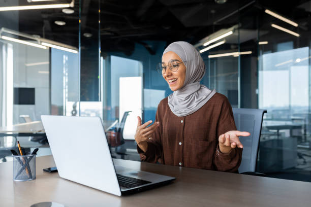 Successful and happy woman inside office at workplace, businesswoman boss in hijab talking on video call using laptop sitting at table, muslim woman joyfully gesturing to colleagues in online meeting Successful and happy woman inside office at workplace, businesswoman boss in hijab talking on video call using laptop sitting at table muslim woman joyfully gesturing to colleagues in online meeting interrogation woman stock pictures, royalty-free photos & images