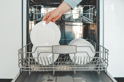 Female hand loading dished, empty out or unloading dishwasher with utensils. Kitchen appliances, lifestyle view. Woman puts a plate in the dishwasher or takes from it. Housewife does her housework.