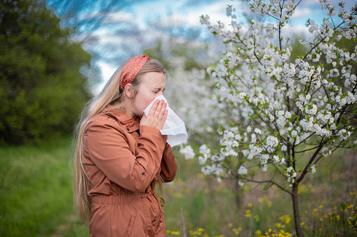 Young woman with pollen allergy holding tissue and blowing her nose outdoors at springtime.