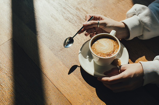 A mug of aromatic coffee in the hands of a woman in a white sweater in a cafe with soft lighting.