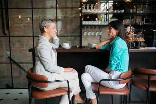Side view of two businesswomen sitting at cafe counter having a casual talk. Female professionals meeting in a coffee shop.