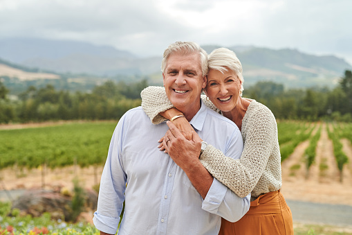 Mature woman smiling and hugging cheerful husband from behind in vineyard, looking at camera