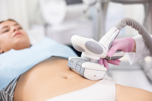 Young woman getting ultrasound cavitation body contouring treatment, anti-cellulite therapy, vacuum massage from unrecognizable cosmetologist. Hardware cosmetology for body sculpting