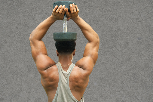 istock Dumbbell for back and shoulder workout:  A strong African male standing and holding a dumbbell up his head in a solid gray background. 1483151975